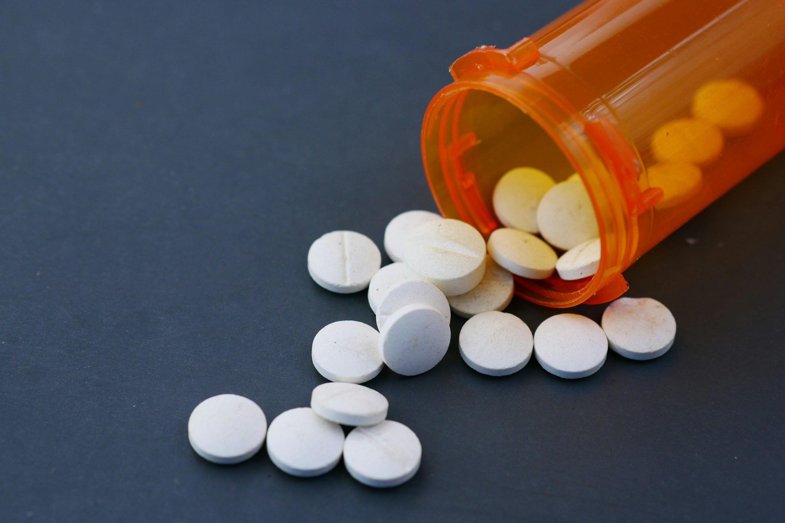 Close-up of white pills scattered from an orange prescription bottle on a gray surface.